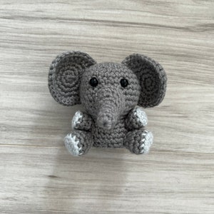 May include: A handmade gray crochet elephant with large ears, a long trunk, and small black eyes. The elephant's feet have white and gray accents. This soft toy is perfect for a baby or child.