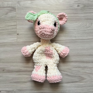 May include: A handmade crochet cow plushie in white, pink, and green. The cow has a white body with pink spots and accents on the hooves, ears, and snout. A green hat sits atop the cow's head. The plushie has black safety eyes.