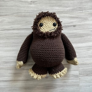 May include: A crocheted plush toy shaped like a creature with a round dark brown body, long arms, and short legs. The face is light beige with a shaggy dark brown border, black plastic eyes, and a small embroidered mouth. The hands and feet are beige.