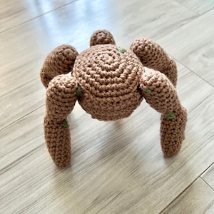 May include: A handmade crochet spider toy in shades of brown, with a rounded body and eight articulated legs. The spider has a spiral pattern on its back and small green accents. The toy is designed to sit on a surface.