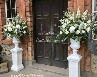 May include: Two white urns filled with white flowers and greenery, each atop a white pedestal. The urns are placed on either side of a dark wooden door. The text "Jackon Customization" is visible on the door. The size is 55cm.