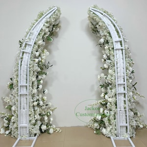 May include: Two white metal wedding arches, each adorned with abundant white flowers and greenery. The arches have a curved top and a rectangular base, creating a visually appealing backdrop for ceremonies or events. The arches are approximately 7 feet tall.