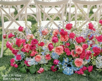 May include: A vibrant floral arrangement featuring pink and red roses, blue hydrangeas, and white flowers. The arrangement is 3.3ft/100cm long, 1.3ft/40cm wide, and 1.8ft/55cm high, placed in front of white chairs on a grassy lawn.