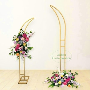 Könnte beinhalten: Zwei goldene Metallbogenständer mit Blumenarrangements. Die Bogenständer sind hoch und schlank mit einer gebogenen Oberseite. Die Blumenarrangements bestehen aus künstlichen Blumen in verschiedenen Farben, darunter rosa, lila, gelb und weiß.