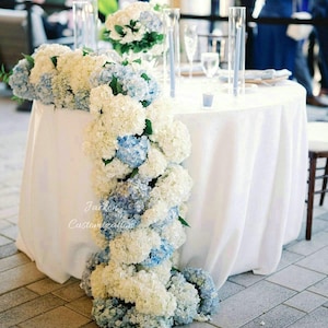 Könnte beinhalten: Ein runder Tisch mit weißer Tischdecke, geschmückt mit einer Kaskade aus weißen und blauen Hortensienblüten. Auf dem Tisch befinden sich auch hohe Glasvasen mit Kerzen und ein kleiner Blumenstrauß. Die Blumengestecke sind etwa 200 cm lang.