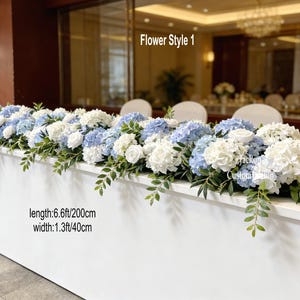 May include: A white table adorned with a floral display of white and pale blue hydrangeas, white roses, and green leaves. The arrangement is 200cm long and 40cm wide. The text "Flower Style 1" is visible.