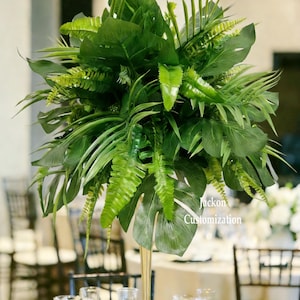 May include: A lush, green artificial floral arrangement, 22 inches (55 cm) tall, featuring a variety of tropical leaves and ferns. The arrangement is displayed on a gold-colored, slender, trumpet-shaped stand, suitable for table centerpieces. The text "Flower Style 1" is visible.