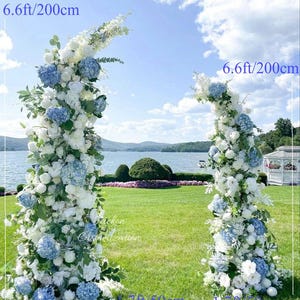 May include: Two floral archways, each 6.6ft (200cm) tall and 1.7ft (50cm) wide, adorned with white and blue flowers. The arches are set on a green lawn with a lake and gazebo in the background, under a blue sky.