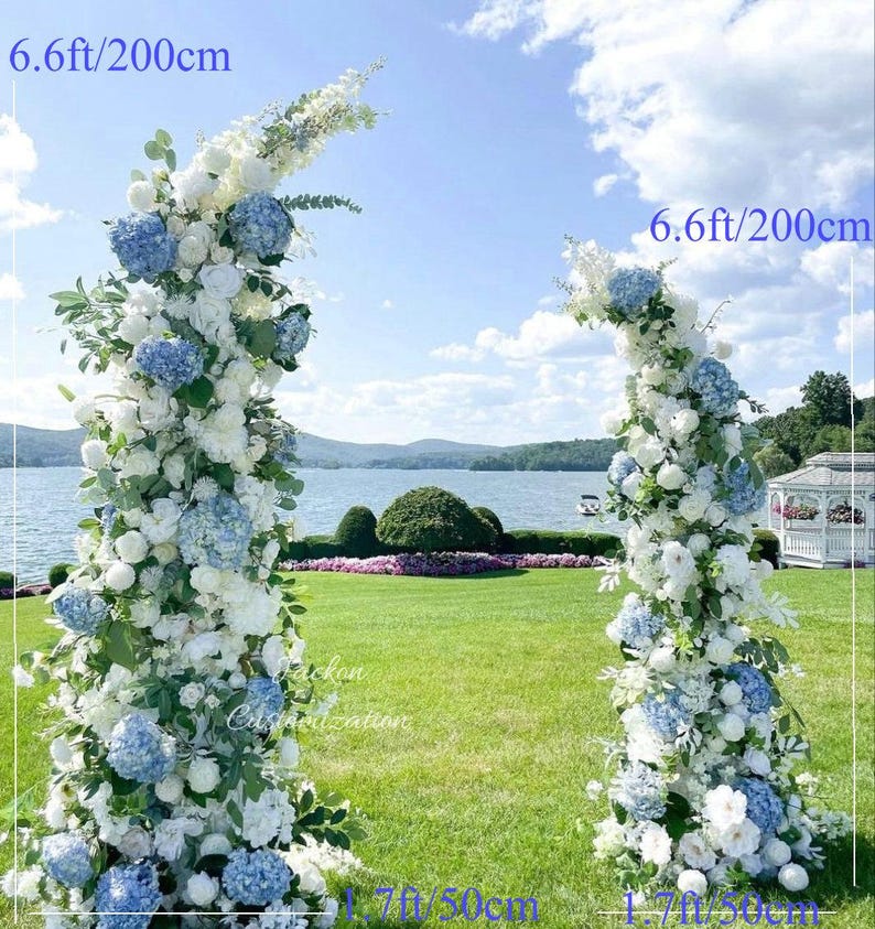 May include: Two floral archways, each 6.6ft (200cm) tall and 1.7ft (50cm) wide, adorned with white and blue flowers. The arches are set on a green lawn with a lake and gazebo in the background, under a blue sky.