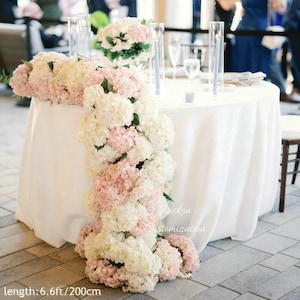 Pode incluir: Uma mesa redonda coberta com uma toalha de mesa branca, adornada com flores de hortênsia brancas e rosas em cascata. O arranjo inclui um centro de mesa floral e dois castiçais altos de vidro. O texto "Jackon Customization" é visível. O comprimento é de 200 cm.