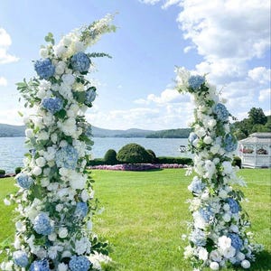 May include: Two floral archways, adorned with white and blue hydrangeas and other white flowers, create a picturesque wedding ceremony setting. The arches frame a scenic view of a lake, mountains, and a bright blue sky with fluffy clouds.