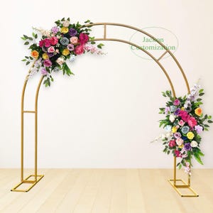 May include: A gold metal archway decorated with colorful artificial flowers. The archway is made of two curved metal pieces that meet at the top. The flowers are arranged in a cluster on the left side of the archway and a smaller cluster on the right side. The archway is standing on a wooden floor.