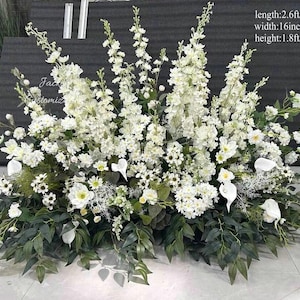 May include: A large white floral arrangement featuring various flowers and foliage. The arrangement measures 80cm long, 40cm wide, and 55cm high. The text "Jack Customic" is visible. Includes calla lilies and daisies.