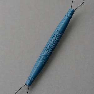 May include: A blue pottery loop tool with the text "YK STUDIO" printed on the handle. The tool has a wire loop on each end, designed for sculpting and trimming clay. The tool is set against a gray background.
