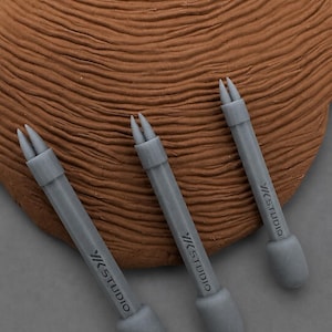 May include: Three gray YK STUDIO tools with a pointed tip and rounded base are arranged on a textured brown surface. The tools are angled, with the YK STUDIO logo visible on the shaft of each tool.