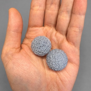 May include: Two small, textured, gray spheres rest in an open palm. The spheres have a detailed, honeycomb-like surface. The background is a solid, neutral gray.