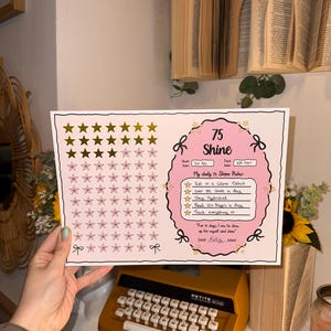 May include: A white and pink 75 Shine tracker with gold stars, bows, and text. The tracker includes daily rules: eat in a calorie deficit, over 8k steps, stay hydrated, read 10+ pages, and track everything. A vintage orange typewriter is in the foreground.