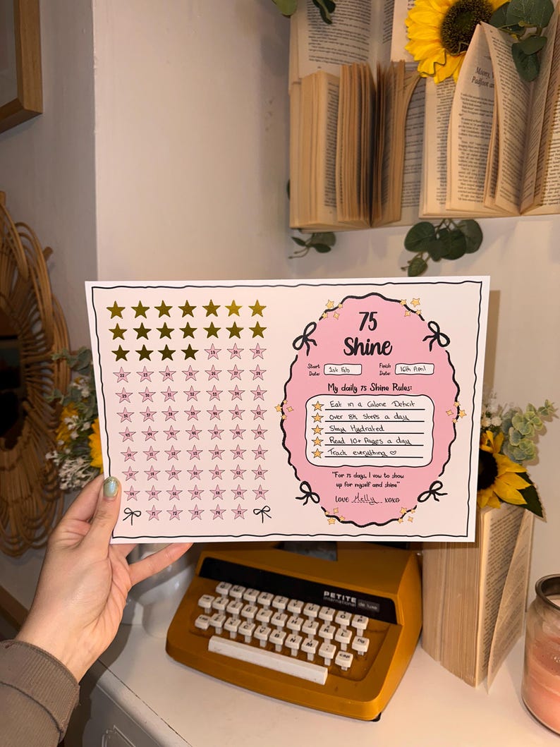 May include: A white and pink 75 Shine tracker with gold stars, bows, and text. The tracker includes daily rules: eat in a calorie deficit, over 8k steps, stay hydrated, read 10+ pages, and track everything. A vintage orange typewriter is in the foreground.