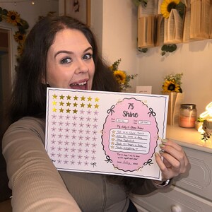 May include: A pink and white checklist with gold stars and the text "75 Shine" is held up by a person. The checklist includes daily goals such as "Eat in a Calorie Deficit" and "Read 100 Pages a day."