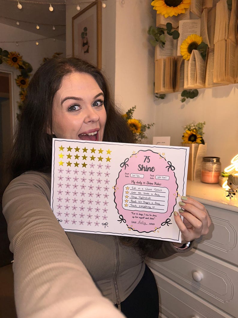 May include: A pink and white checklist with gold stars and the text "75 Shine" is held up by a person. The checklist includes daily goals such as "Eat in a Calorie Deficit" and "Read 100 Pages a day."