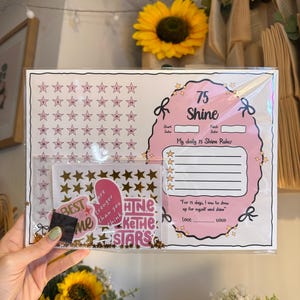 May include: A pink and white 75 Shine daily rules chart with star-shaped stickers. The chart includes space for daily rules and a motivational quote. Gold star stickers and the words "BEST ME" are also included.