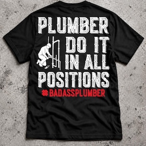 以下が含まれることがあります： 黒の半袖Tシャツで、白い文字で「PLUMBER DO IT IN ALL POSITIONS」と書かれ、配管作業をする人物のグラフィックが描かれています。テキストの下には赤いハッシュタグ記号と、赤い文字で「BADASSPLUMBER」と記されています。