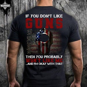 May include: A black short-sleeved t-shirt featuring a graphic print on the back. The text reads IF YOU DON'T LIKE GUNS THEN YOU PROBABLY WON'T LIKE ME...AND I'M OKAY WITH THAT. A central image shows a gun barrel overlaying a United States flag.