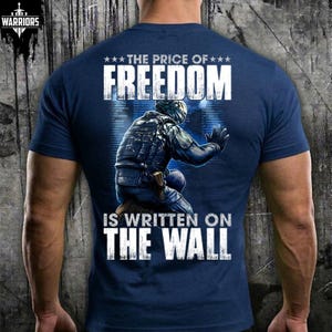 May include: A navy blue short-sleeve t-shirt featuring a graphic on the back of a tactical soldier kneeling against a wall. The text reads THE PRICE OF FREEDOM IS WRITTEN ON THE WALL. The design includes a small logo in the top left corner.