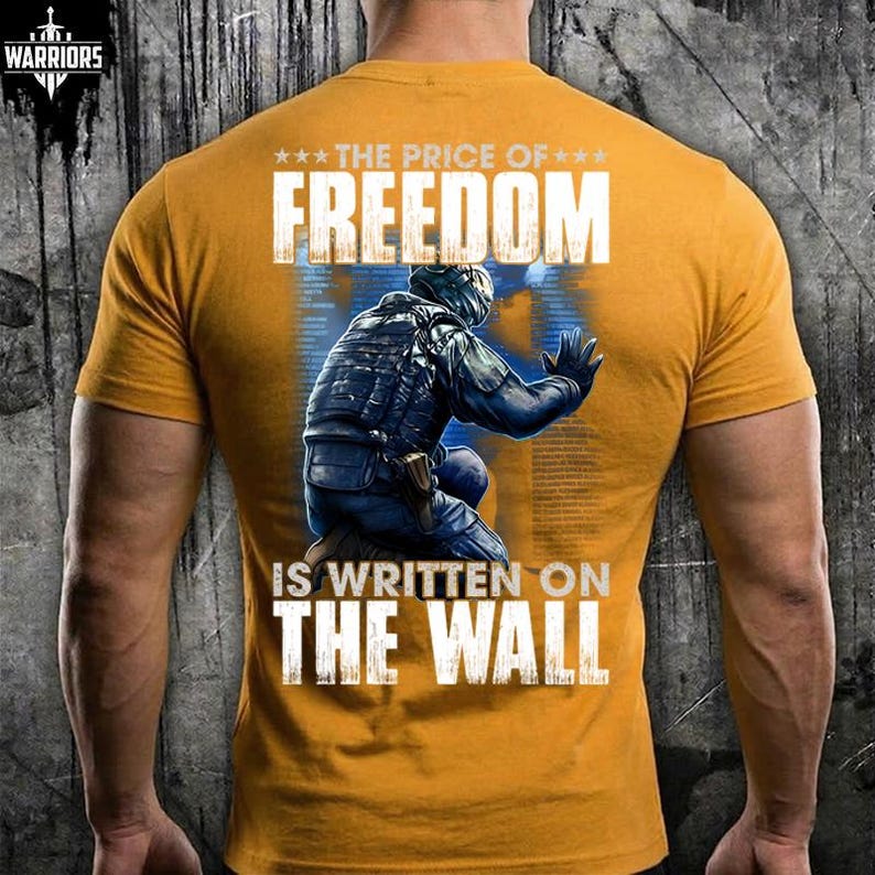 May include: A mustard yellow short-sleeve t-shirt featuring a graphic on the back of a tactical soldier kneeling and touching a wall. The text reads THE PRICE OF FREEDOM IS WRITTEN ON THE WALL in white capital letters with three stars above.