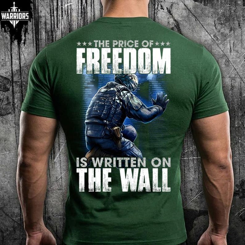 May include: A forest green short-sleeve t-shirt featuring a graphic on the back of a tactical soldier kneeling against a blue digital wall. The text reads THE PRICE OF FREEDOM IS WRITTEN ON THE WALL in white capital letters with three stars.