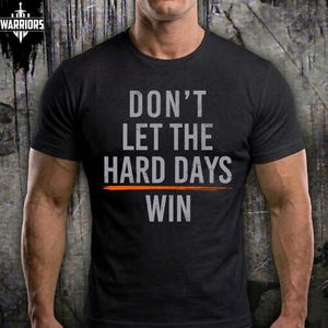 Puede incluir: Una camiseta negra de manga corta con el texto DON'T LET THE HARD DAYS WIN en letras grises desgastadas, una línea horizontal naranja que separa las palabras HARD DAYS y WIN, y un pequeño logotipo en la esquina superior izquierda.