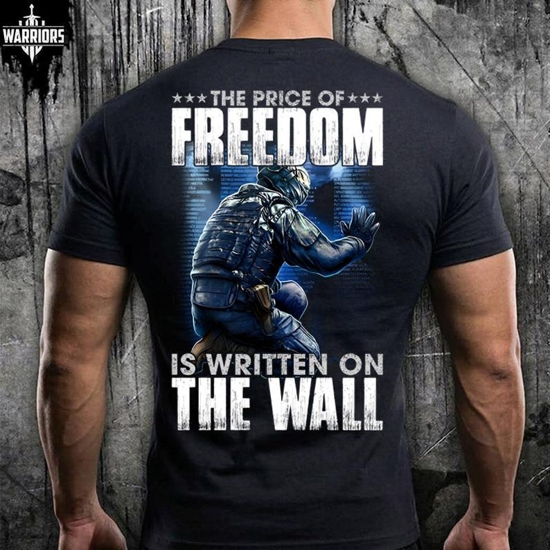 May include: A black short-sleeved t-shirt featuring a graphic on the back of a soldier in tactical gear kneeling and touching a wall. The text reads THE PRICE OF FREEDOM IS WRITTEN ON THE WALL. A small logo in the top left corner says WARRIORS.