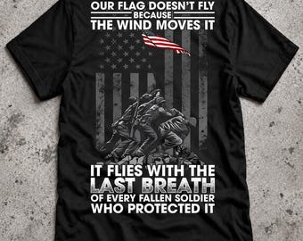 May include: A black t-shirt featuring a graphic of soldiers raising an American flag. The text reads: OUR FLAG DOESN'T FLY BECAUSE THE WIND MOVES IT IT FLIES WITH THE LAST BREATH OF EVERY FALLEN SOLDIER WHO PROTECTED IT. The design includes a flag motif.