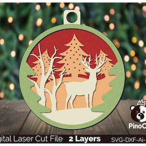 May include: A layered, laser-cut Christmas ornament featuring a deer standing in a snowy forest scene. The ornament is red, green, and white with a brown deer silhouette. The ornament has a hole at the top for hanging.