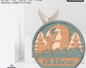 May include: A round wooden sign with a light blue background and a cutout of the letter "A" surrounded by trees and a deer. The name "Alice" is written below the letter.