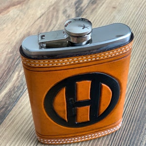 May include: An orange leather flask with a silver top and a black oval with a black letter "H". The flask has white stitching around the edges. The flask is sitting on a wooden surface.