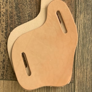 May include: Two pieces of unfinished, light tan leather with a curved shape and rectangular cutouts. The leather has a natural, unpainted appearance, with one piece slightly overlapping the other. The background is a wooden surface.