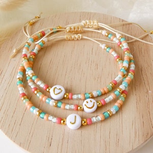 Könnte beinhalten: Drei verstellbare Armbänder aus kleinen Glasperlen in Pfirsich-, Türkis- und Weißtönen, akzentuiert mit goldfarbenen Metallperlen. Jedes Armband hat eine weiße, runde Perle mit einem goldenen Herz, einem Smiley-Gesicht oder dem Buchstaben J.