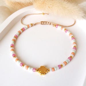 Könnte beinhalten: Ein Perlenarmband mit einem sich wiederholenden Muster aus kleinen rosa, pfirsichfarbenen, weißen und metallisch goldenen Perlen. Ein zentraler, goldfarbener vierblättriger Kleeblatt-Anhänger bildet den Mittelpunkt. Das Armband hat ein verstellbares Band.