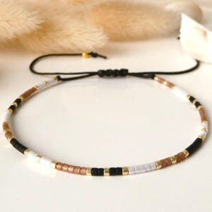 Könnte beinhalten: Ein zartes Armband mit einem sich wiederholenden Muster aus kleinen zylindrischen Perlen in den Farben Mattschwarz, Metallic-Gold, Weiß und Kupfer. Das Armband ist auf einer dünnen schwarzen Kordel mit einem verstellbaren Schiebeknotenverschluss.