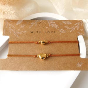 Könnte beinhalten: Zwei Armbänder auf einer braunen rechteckigen Karte mit dem Text WITH LOVE. Jedes Armband besteht aus einer dünnen orangefarbenen Kordel, einem kleinen goldfarbenen Metallherz-Anhänger und zwei winzigen goldfarbenen Perlen neben dem Herz. Die Karte hat weiße florale Linien.
