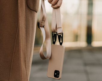 May include: A tan phone case with a matching lanyard. The lanyard is made of a woven fabric and has black metal clips. The phone case is a solid color and has a camera lens on the back.
