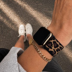 Könnte beinhalten: Eine Smartwatch mit schwarzem Display und goldfarbenem Rahmen, ausgestattet mit einem elastischen Uhrenarmband mit schwarzem und braunem Leopardenmuster. Ein goldfarbenes Metallkettenarmband mit einem kleinen runden Anhänger am selben Handgelenk.