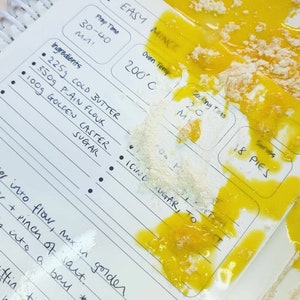 Pu&ograve; includere: Una pagina di ricetta bianca con testo scritto a mano che include Easy Mince, 225g di burro freddo, 350g di farina, 100g di zucchero, 200 gradi Celsius e 18 torte. La pagina &egrave; coperta da liquido giallo brillante e farina bianca.