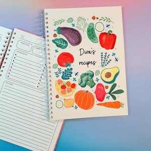 Pu&ograve; includere: Un ricettario con rilegatura a spirale e copertina bianca con illustrazioni colorate di verdure, tra cui melanzana, pomodoro, broccoli, zucca e carota. Il testo "Diwa's recipes" &egrave; stampato al centro. Una pagina aperta mostra i campi per le ricette.