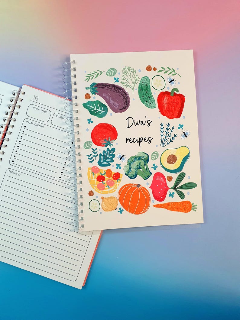 Pu&ograve; includere: Un ricettario con rilegatura a spirale e copertina bianca con illustrazioni colorate di verdure, tra cui melanzana, pomodoro, broccoli, zucca e carota. Il testo "Diwa's recipes" &egrave; stampato al centro. Una pagina aperta mostra i campi per le ricette.