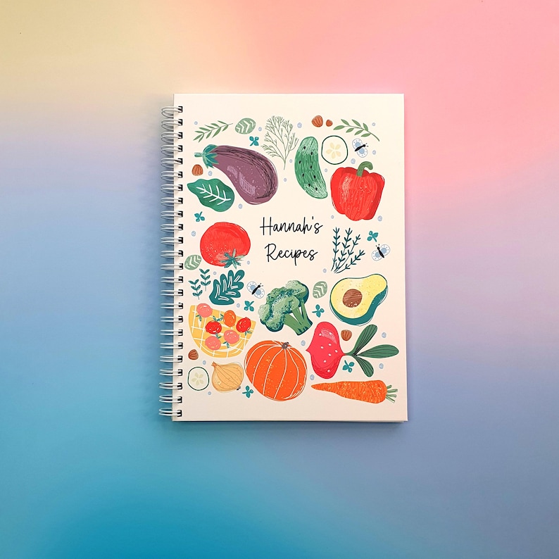 Pu&ograve; includere: Un quaderno a spirale bianco con una colorata illustrazione di varie verdure, tra cui una melanzana, un peperone rosso, broccoli, una zucca, una carota e un avocado. Il testo "Hannah's Recipes" &egrave; stampato al centro in corsivo nero.