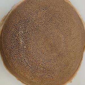 May include: A round, flat injera bread with a light brown color and a distinct porous, sponge-like texture across its entire surface. The edges are slightly raised and uneven, showing the thin, flexible nature of this fermented flatbread.