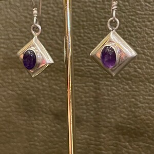 May include: A pair of silver earrings with a square shape and a purple amethyst gemstone in the center.