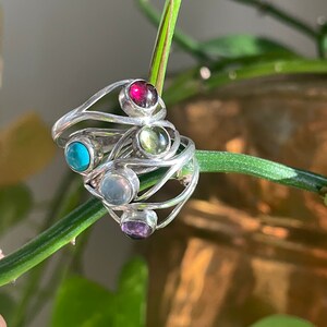 Puede incluir: Un anillo de plata con cinco piedras preciosas de diferentes colores. El anillo está hecho de alambre de plata trenzado y presenta una piedra roja, verde, azul, blanca y morada.